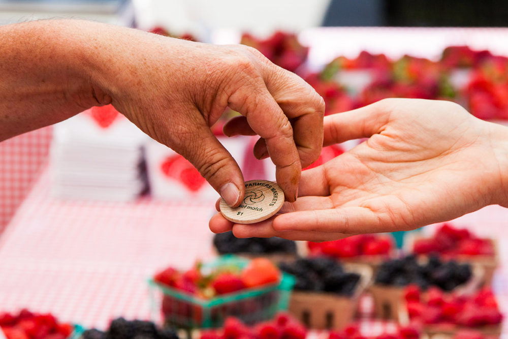 Hands exchanging a CalFresh farmers market token with berries in the background