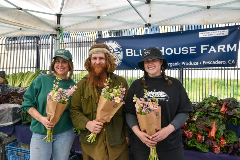 Three people hold flower bouquets at Blue House Farm's stand at Foodwise's Mission Community Market in San Francisco