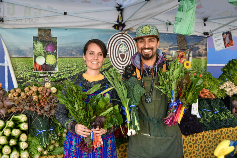 Maria and Rudy hold leafy vegetables and pose at Green Thumb Farms' stand at Foodwise's farmers market in San Francisco