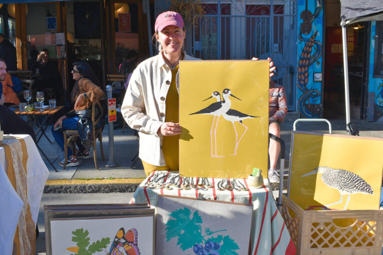 Photo of artist Jen Kindell holding up a poster with two black and white birds on a yellow background at Foodwise's Mission Community Market in San Francisco