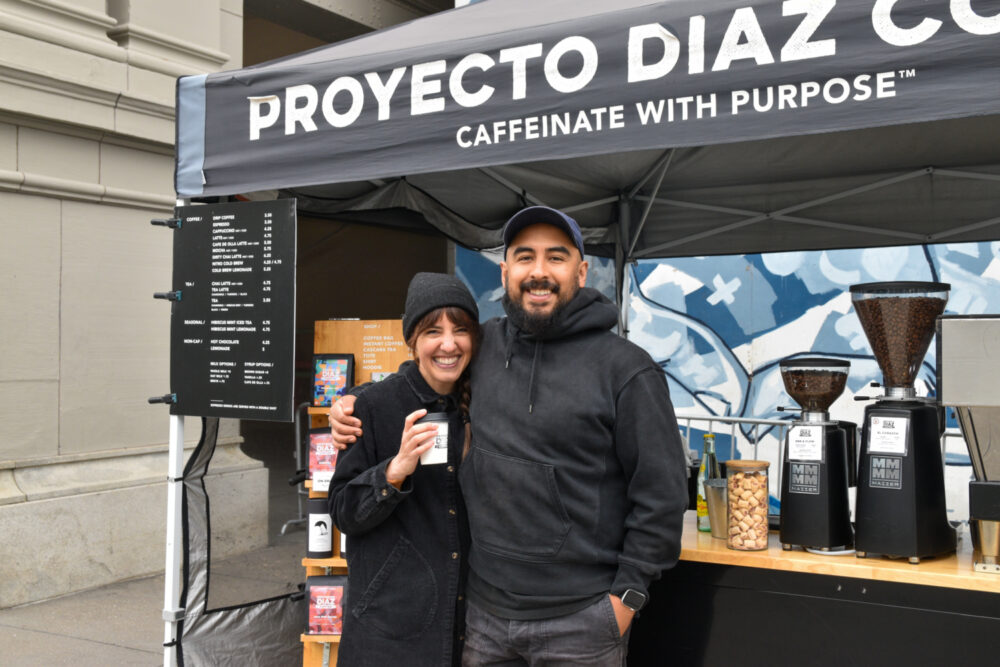 Two people, Hannah and Fernando, hold a cup of coffee in front of Proyecto Diaz Coffee's tent at the Ferry Plaza Farmers Market