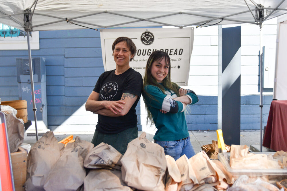 Two people pose back-to-back at Fox and Lion's stand at Foodwise's Mission Community Market in San Francisco