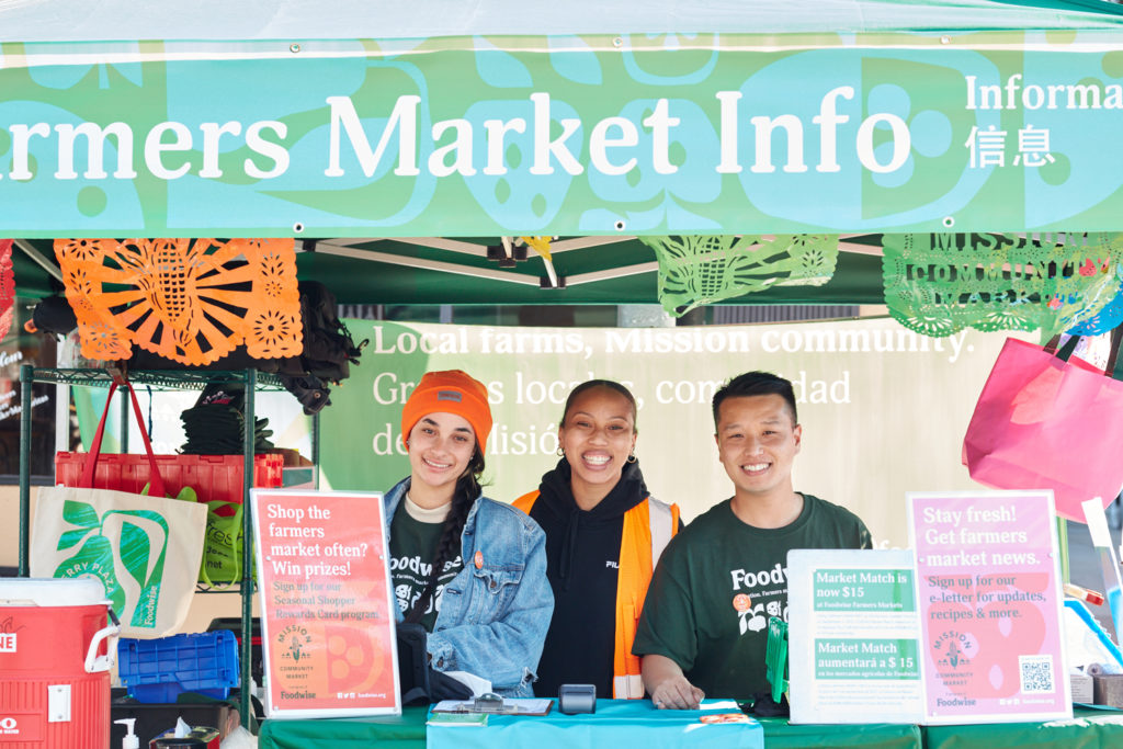 Three Foodwise staff members at the Farmers Market Info Booth, including Deven Okry and Tommy Phung