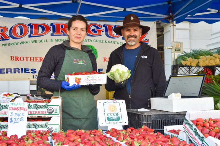 Two people pose at Rodriguez Bros Ranch's stand at the Ferry Plaza Farmers Market. Strawberries are on the table in front of them.