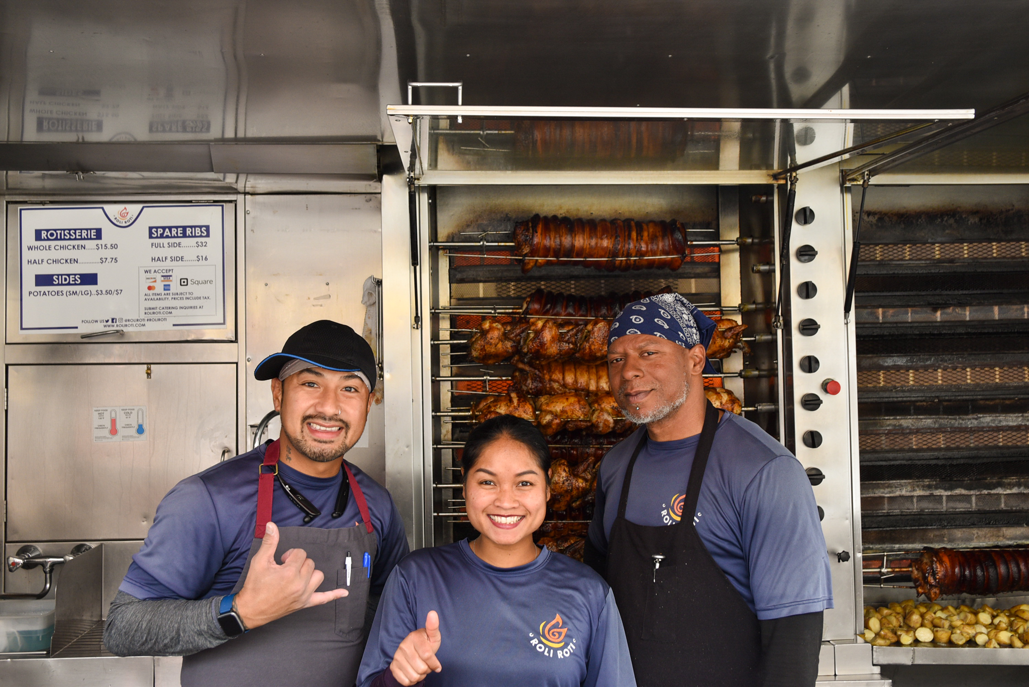 Three people pose in front of the rotisserie at RoliRoti Gourmet Rotisserie's truck at the Ferry Plaza Farmers Market in San Francisco.