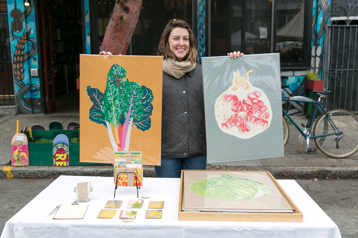 Jen Kindell holds art prints, one of rainbow chard and one of a pomegranate, at her table at the Mission Community Market in San Francisco.