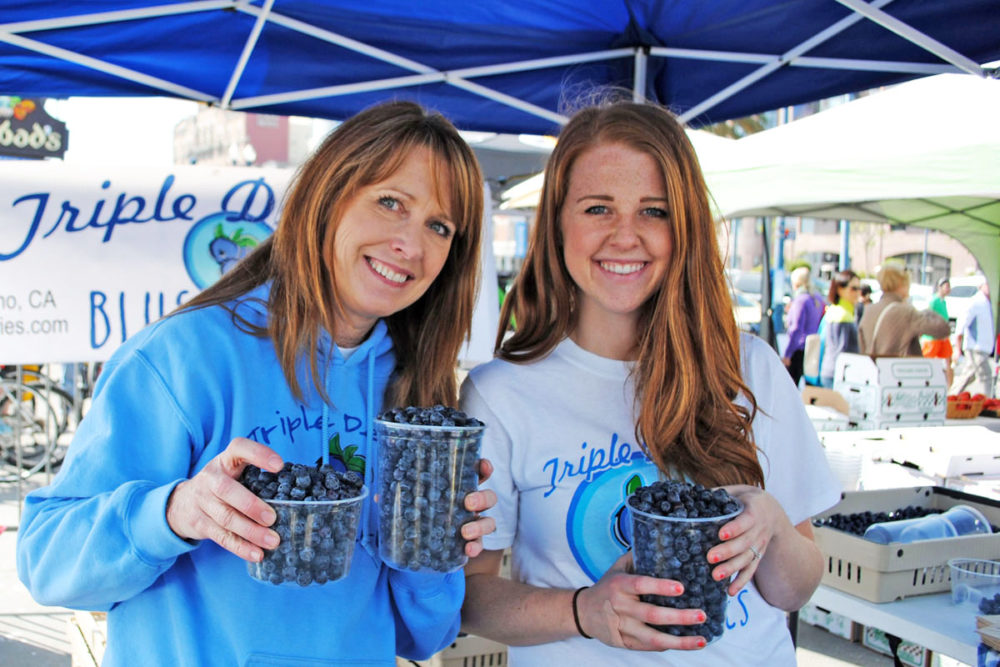 Two people hold blueberries and pose at Triple Dleight Blueberries' stand at the Ferry Plaza Farmers Market in San Francisco.