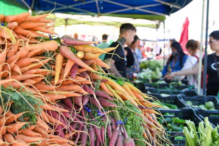 Colorful bunches of carrots at a Ferry Plaza Farmers Market stand with shoppers in the background