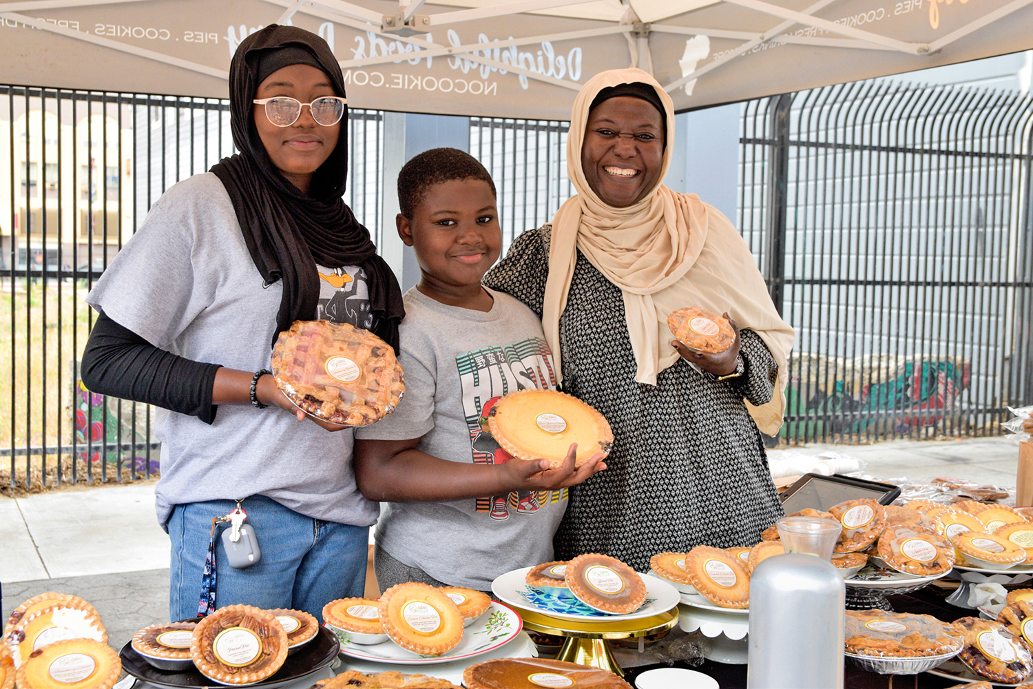 Three people, including Rafia Sabir, hold pies and pose at Delightful Foods' stand at the Mission Community Market in San Francisco.
