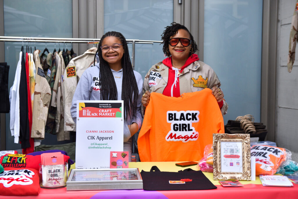 Two people pose with clothing and accessories at CIK Apparel's stand at Foodwise's Pop-Ups on the Plaza event at the Ferry Plaza Farmers Market in San Francisco