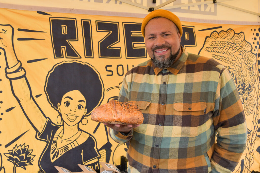 Man holds a loaf of sourdough bread in front of a graphic banner that says "Rize Up"