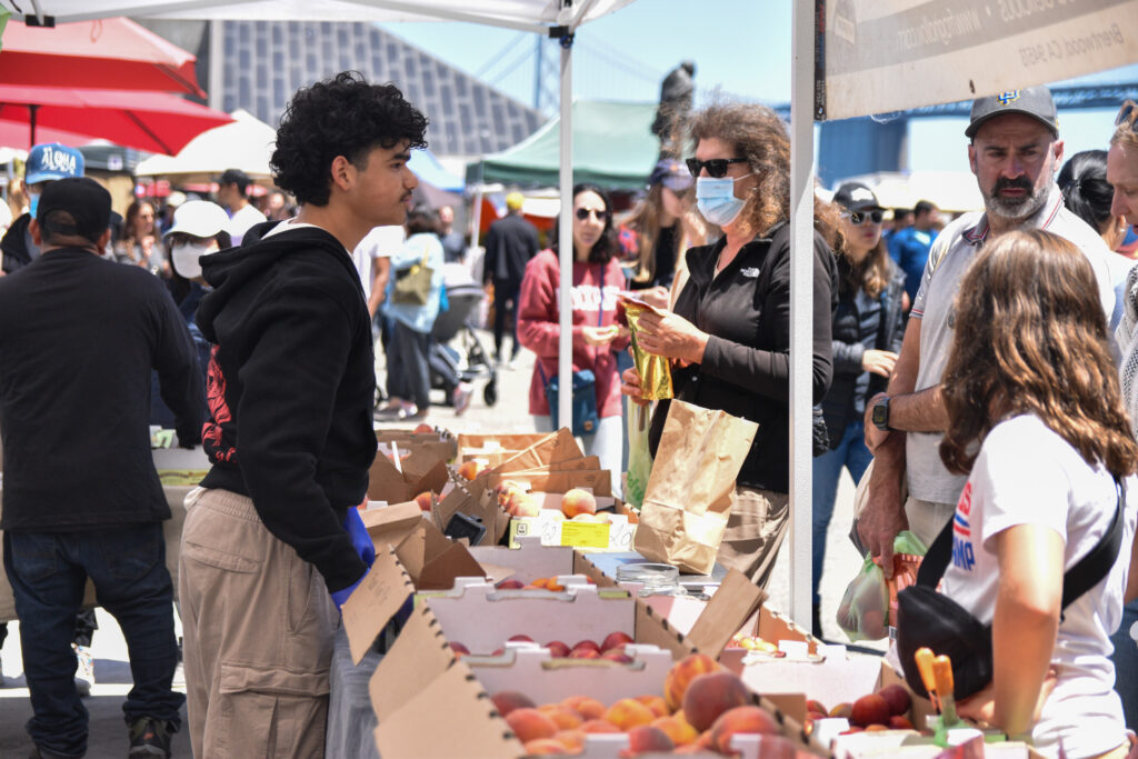 Foodwise Teen, Henry, leans over a table full of stone fruit to talk to shoppers at the Ferry Plaza Farmers Market