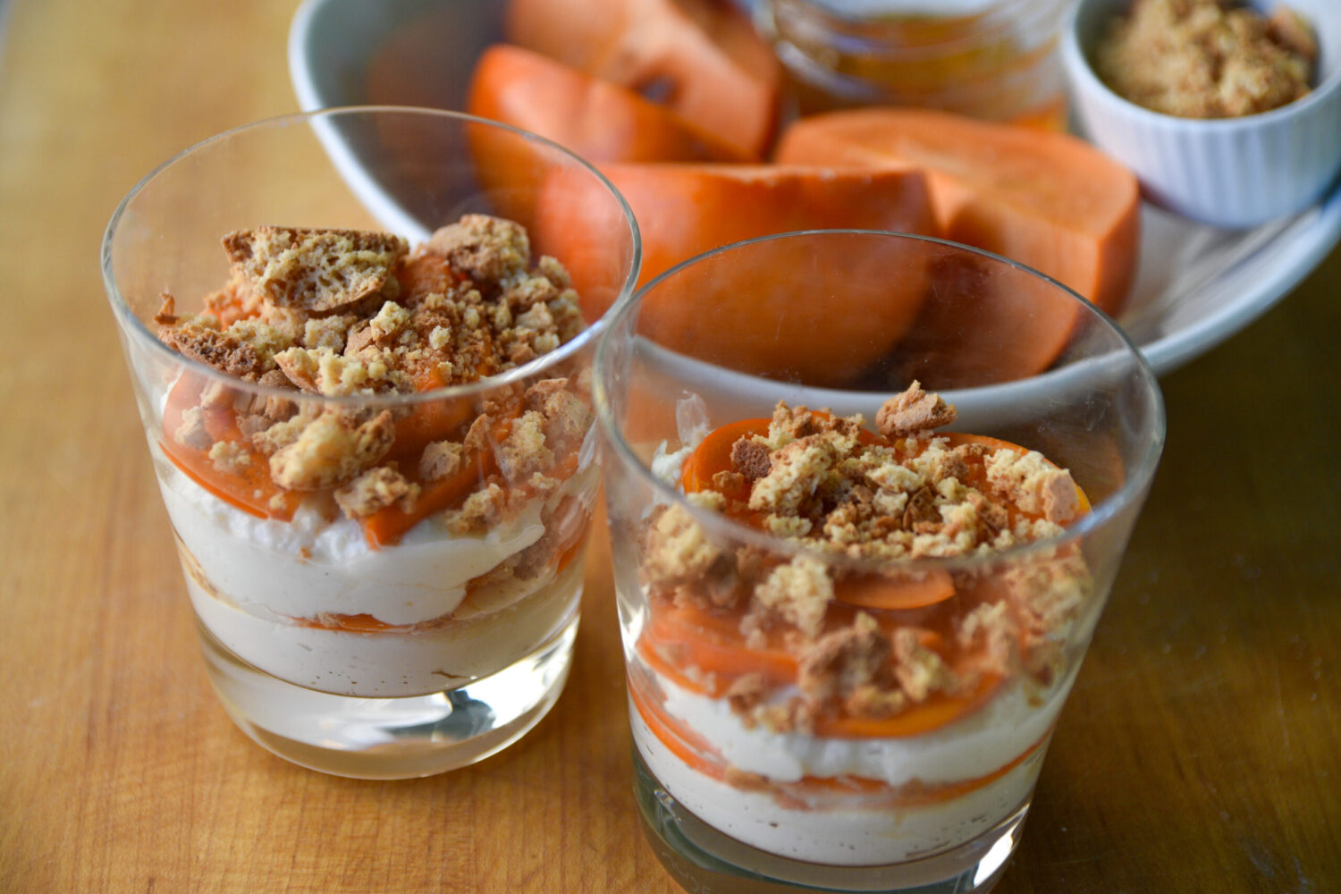 Ricotta Trifle, topped with persimmon slices and crumbled cookies, served in clear cups on a wooden cutting board.