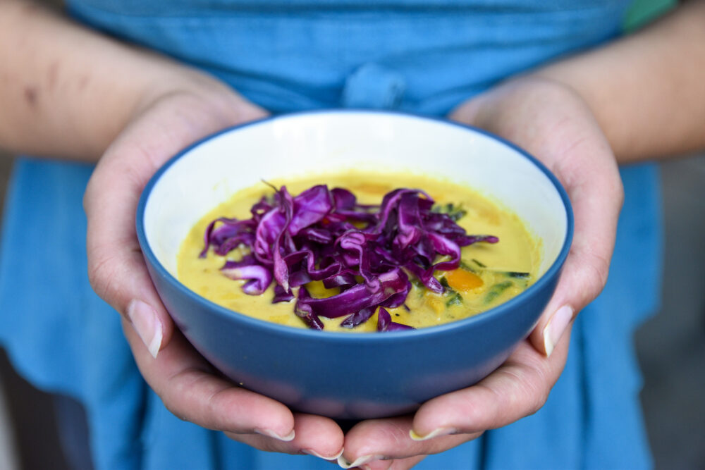 Two hands hold a bowl of yellow winter squash curry, topped with pickled purple cabbage pieces, served in a navy blue bowl.