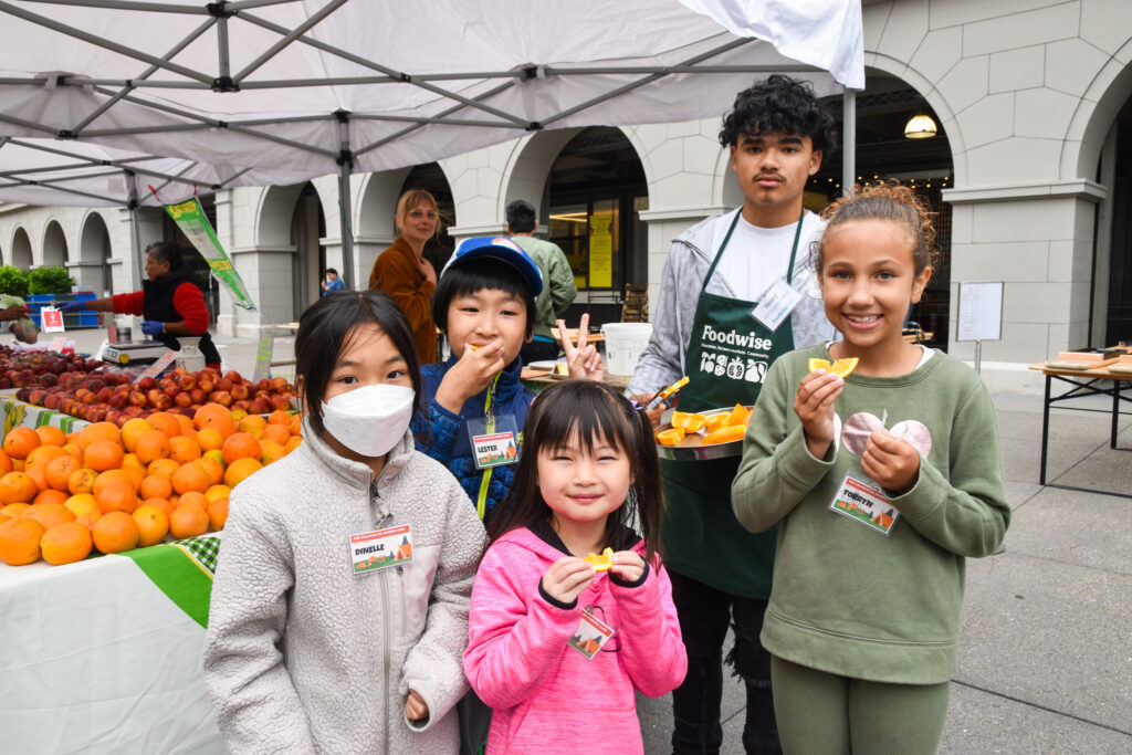 Foodwise Teen, Henry, poses with a group of Foodwise Kids visiting the Ferry Plaza Farmers Market. Tables of citrus and other fruit are in the background.