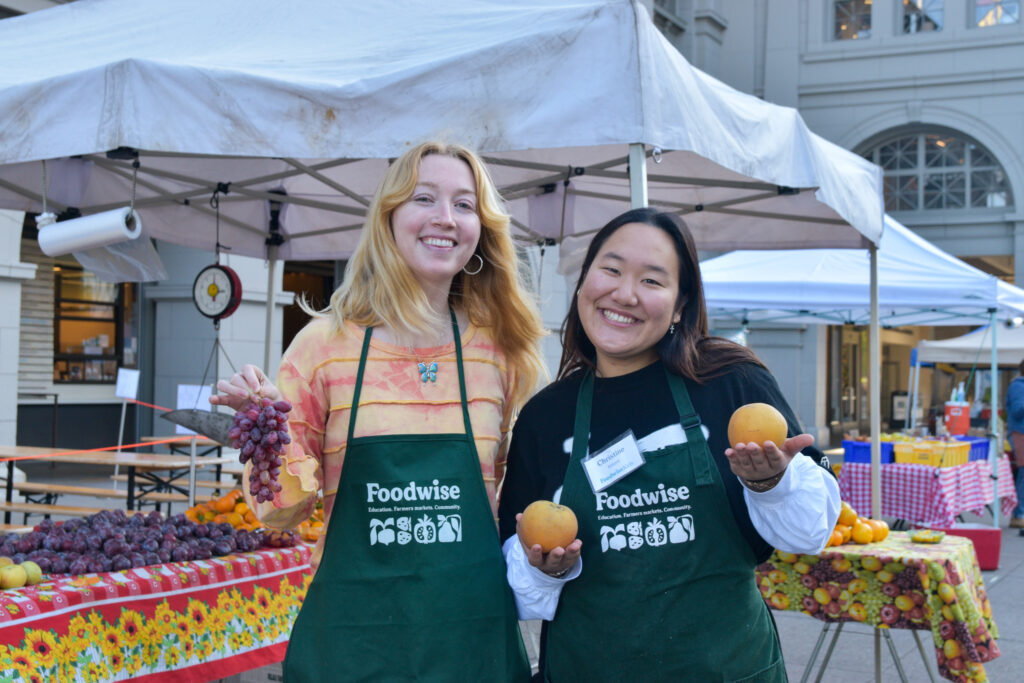 Foodwise Kids interns Heidi Ford and Christine Kim hold fruits in front of a stand at the Ferry Plaza Farmers Market in San Francisco.