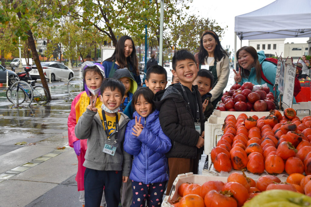 Foodwise volunteers, Kat and Yuki, pose with a group of Foodwise Kids next to a farmers market stand full of persimmons.
