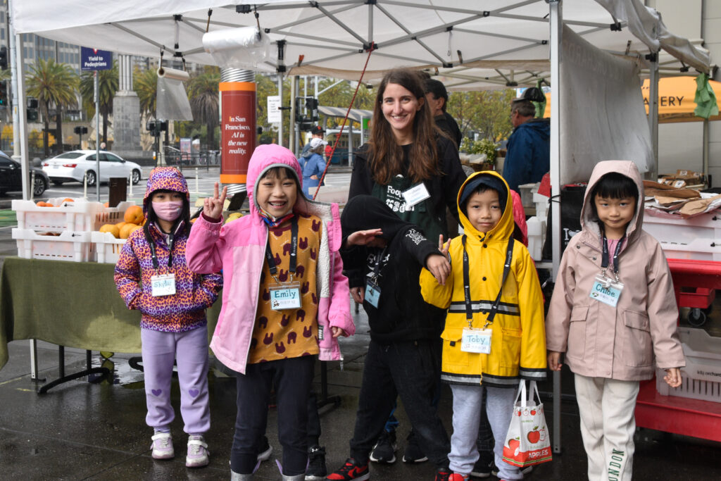 Foodwise volunteer, Stephanie, poses with a group of Foodwise Kids in front of a farm stand at the Ferry Plaza Farmers Market.