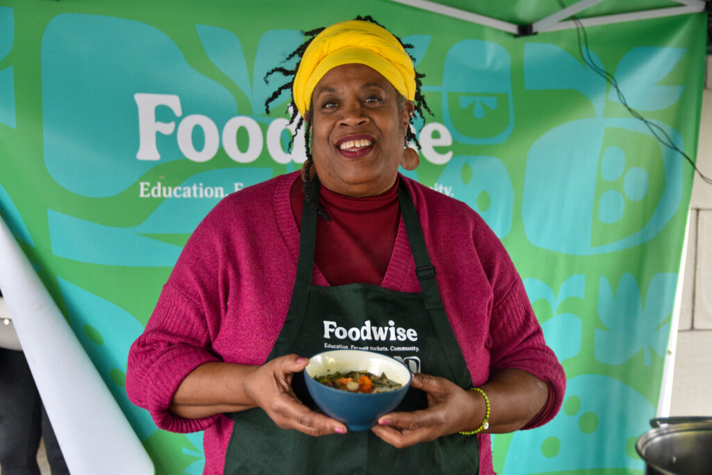 Wanda Blake holds a bowl of food and poses in front of a teal and green backdrop at the Foodwise Classroom at the Ferry Plaza Farmers Market in San Francisco.