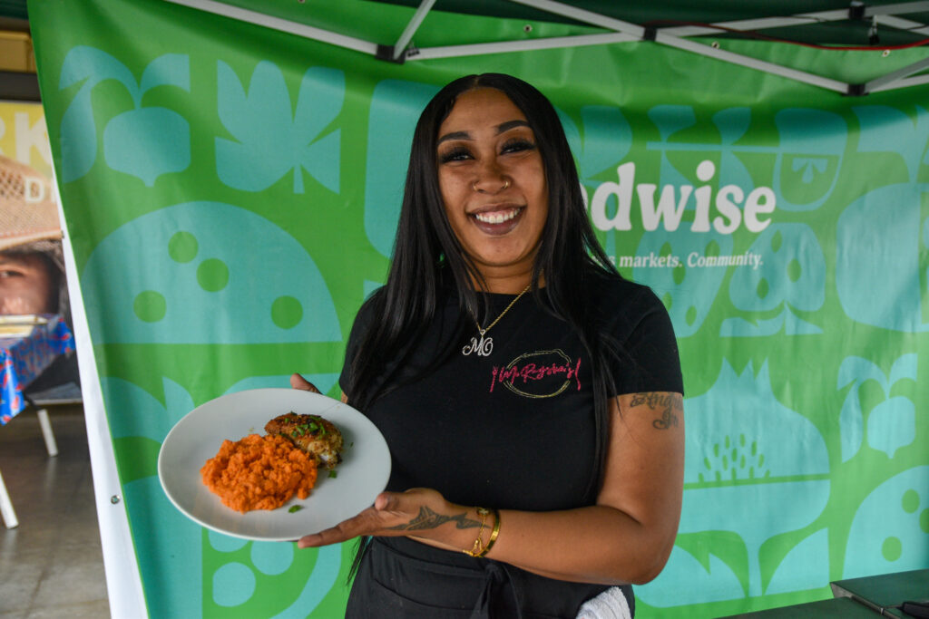 Mo'Raysha Pouoa poses in front of a teal and green background, holding a plate of food, at the Foodwise Classroom at the Ferry Plaza Farmers Market in San Francisco.