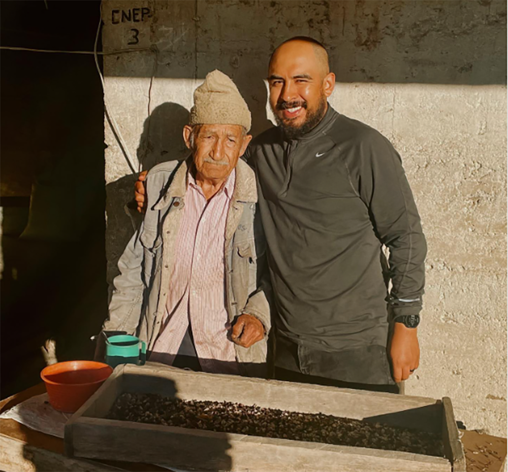Juan and Fernando pose in front of a tray of coffee beans.