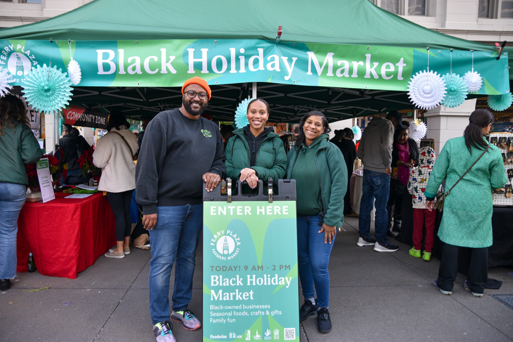 In The Black and Foodwise team members in front of a tent labeled "Black Holiday Market" at Foodwise's Ferry Plaza Farmers Market in San Francisco.