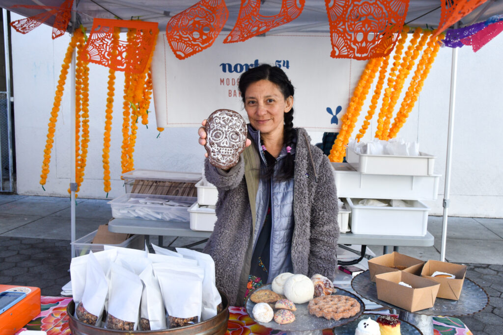 Raquel Goldman holds up a skull cookie at Norte54's stand at Foodwise's Mission Community Market in San Francisco, as strands of carnations blow in the wind in the background