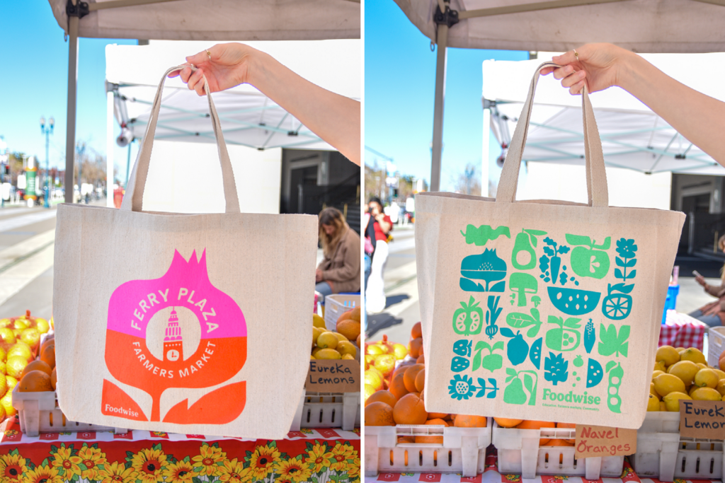 Foodwise tote bags are held up in front of a table holding citrus