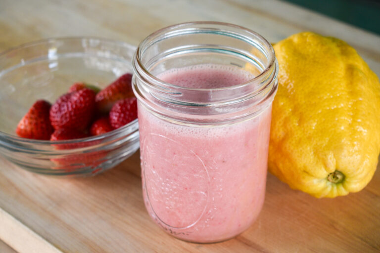 A pink smoothie in a mason jar sits between a bowl of strawberries and a lemon