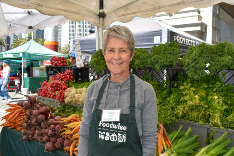 Photo of someone smiling in front of a farmers market stand full of vegetables