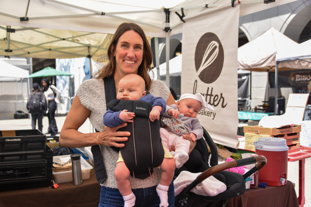 A woman holds two twin babies at the farmers market