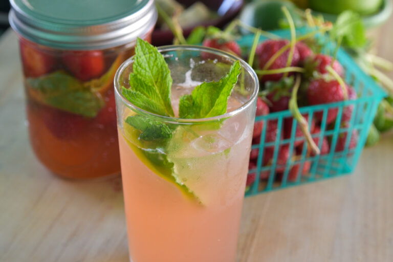 Photo of an iced beverage, with a jar of shrub and a basket of stawberries in the background