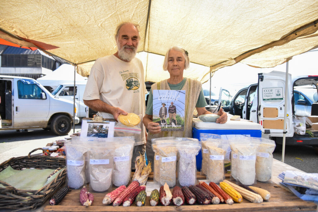 Two people pose at Tierra Vegetables' stand at the Ferry Plaza Farmers Market with heirloom corn, tortillas, and corn products