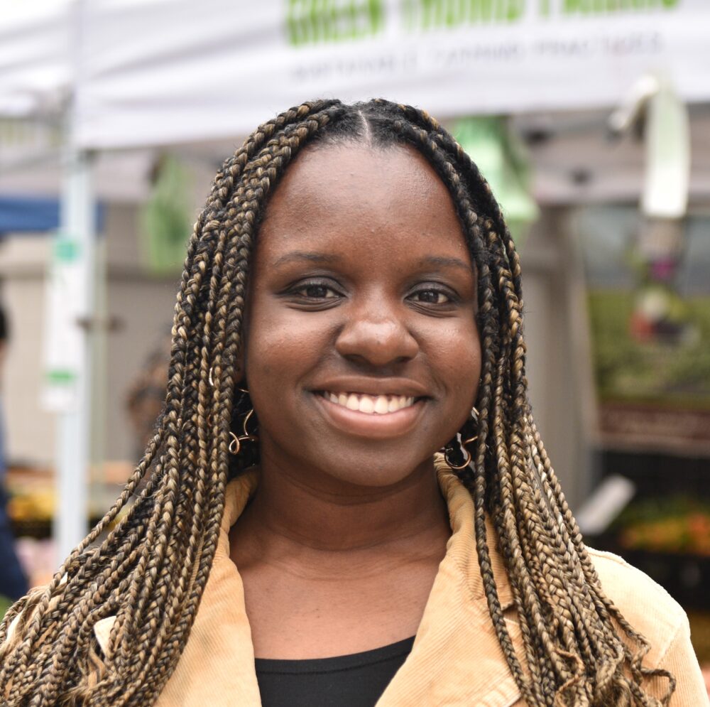 A smiling woman with blonde braids, a beige blazer, and a black shirt.