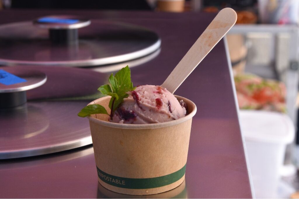 A compostable cup holding a scoop of pink plum gelato with a mint leaf on top. A wooden spoon sticks out of the gelato