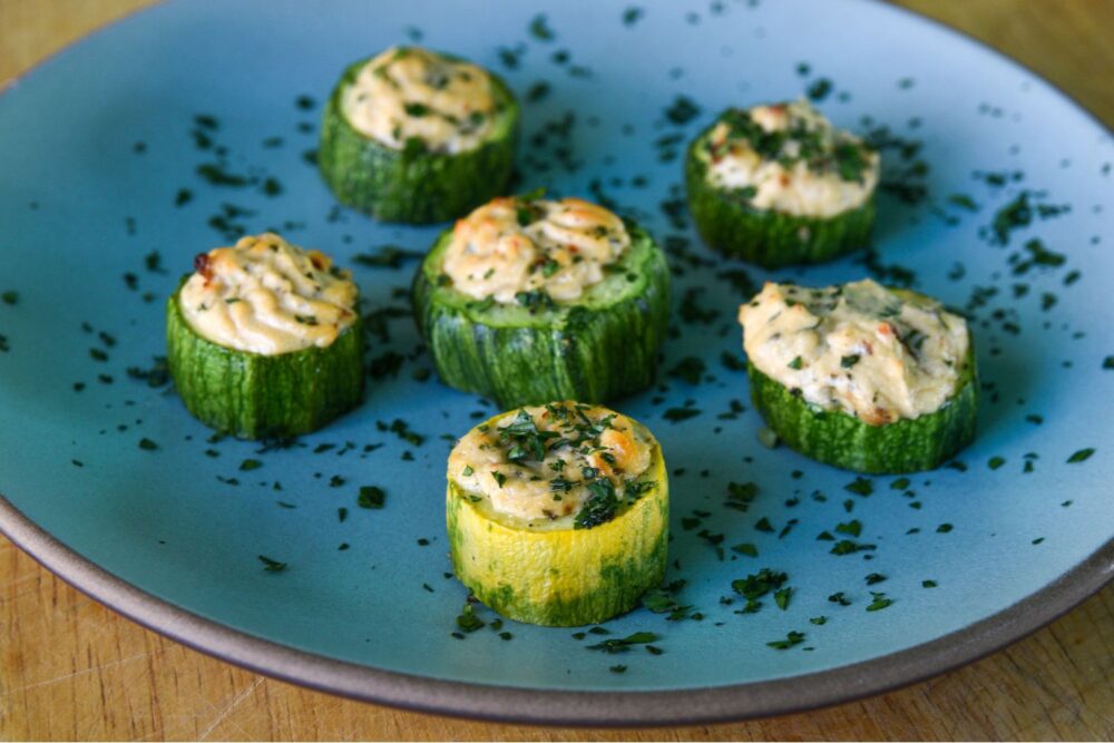 Six zucchini cups on a blue plate. The zucchini cups are stuffed with cheese, and tomatoes, and sprinkled with herbs.